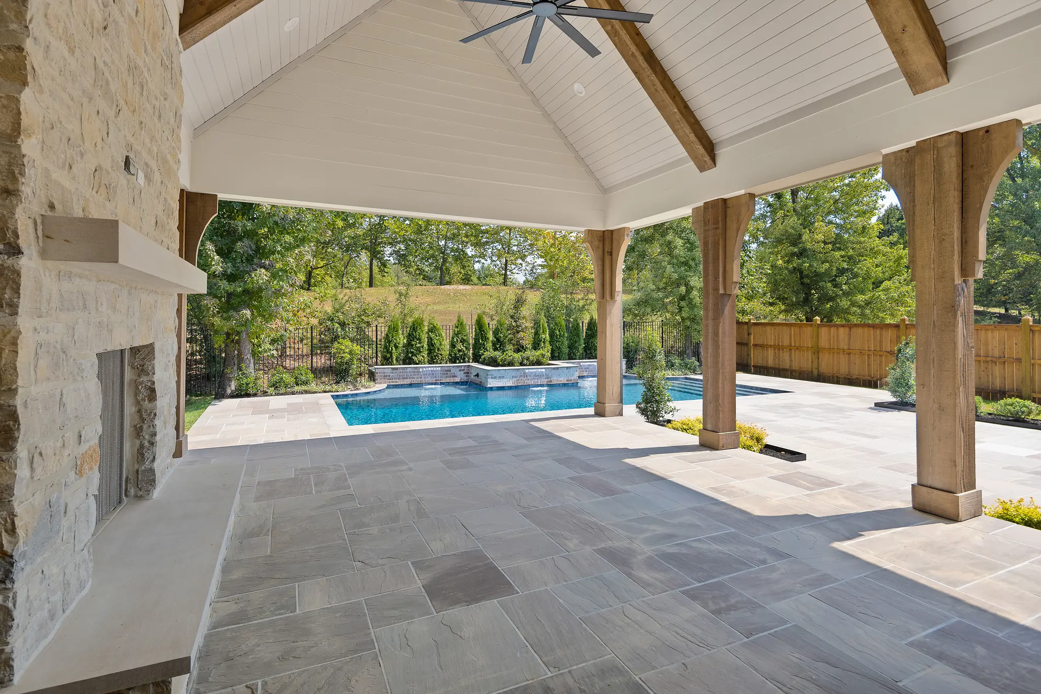 Outdoor Living space with vaulted ceilings, stone flooring, cedar beams and posts, and stone fireplace overlooking vibrant backyard with pool