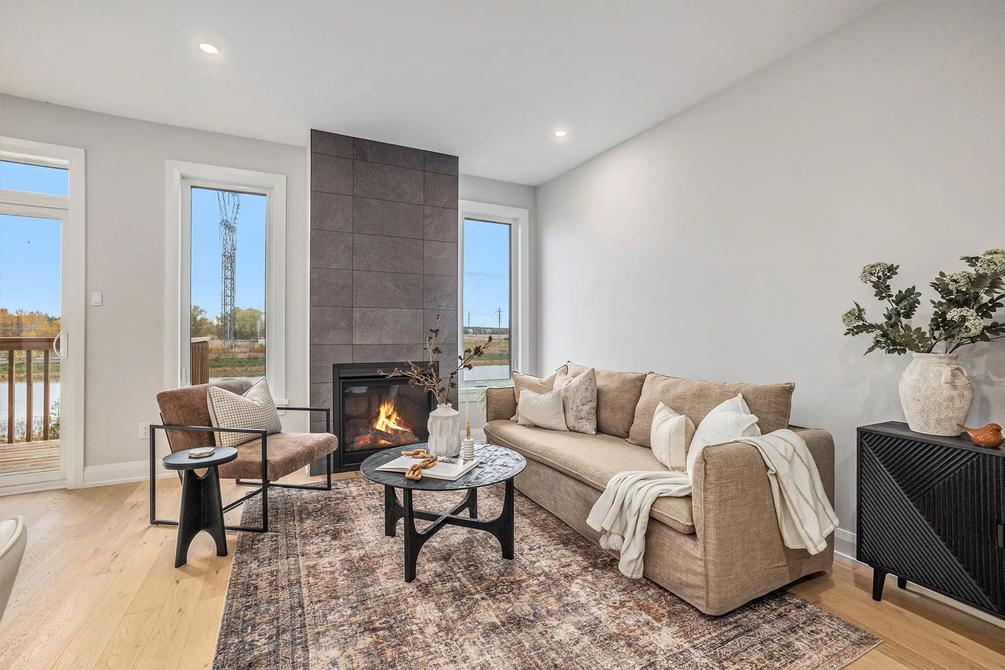Modern living room with fireplace, beige sofa, patterned rug, and large windows